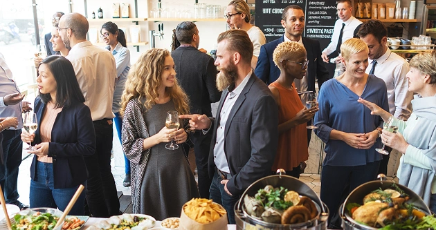 In een stijlvolle ruimte vindt een gezellig samenzijn plaats, waar allerlei mensen met een drankje in de hand een praatje maken, omringd door een smakelijk buffet.
