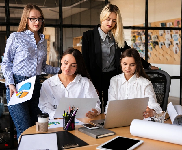 Vier vrouwen werken samen aan een bureau in een modern kantoor, waar ze documenten en een laptop bekijken. De sfeer is geconcentreerd en professioneel.