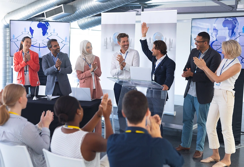 Een diverse groep mensen in zakelijke kleding die klapt en glimlacht tijdens een presentatie met een spreker op het podium, waardoor er een vrolijke en bemoedigende sfeer ontstaat.