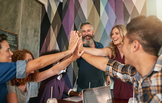 Een diverse groep van vijf mensen in vrijetijdskleding geeft elkaar vrolijk een high-five rond een tafel, met laptops en drankjes. De sfeer is levendig en gezellig.