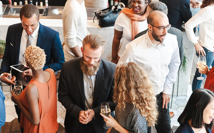 Een diverse groep mensen die gezellig bij elkaar zit tijdens een levendig evenement in een moderne locatie. De aanwezigen glimlachen, hebben een drankje in de hand en zijn in gesprek.