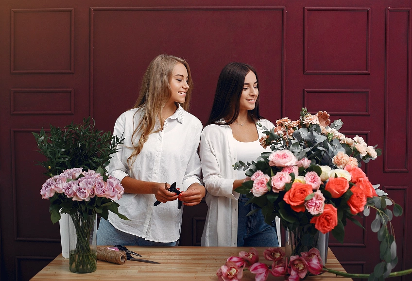 Twee vrouwen in witte blouses schikken kleurrijke bloemen aan een tafel tegen een bordeauxrode muur, terwijl ze glimlachen en met elkaar praten. Het tafereel is levendig en vrolijk.