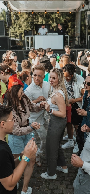 Een bruisend openluchtmuziekfestival met een divers publiek dat uitbundig danst. Op het podium draait een dj, omringd door bomen onder een stralende, heldere hemel.