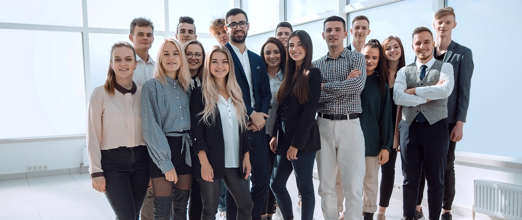 Een groep van 16 jonge professionals staat in een helder verlicht kantoor en glimlacht zelfverzekerd. Ze zijn gekleed in business-casual kleding, wat een gevoel van teamgeest en positiviteit uitstraalt.
