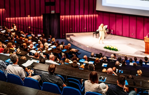 Een spreker houdt een toespraak op het podium in een grote zaal met paarse muren. Het publiek luistert aandachtig vanaf de blauwe tribune, wat de geconcentreerde en betrokken sfeer benadrukt.