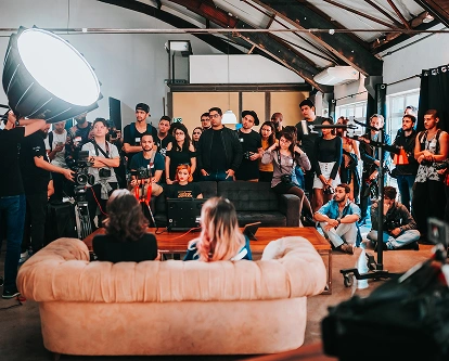 Een grote groep mensen heeft zich in een studio verzameld en kijkt aandachtig naar een presentatie. Felle studiolampen verlichten de ruimte en zorgen voor een sfeer van concentratie en betrokkenheid.