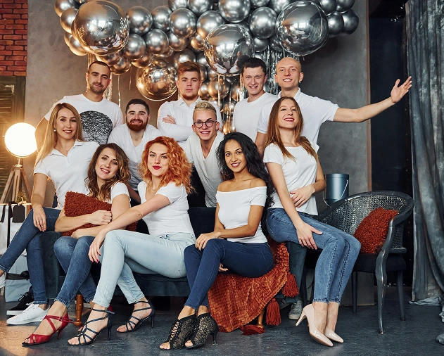 Op de foto is een groep jongeren te zien die samen in een studio poseren voor een achtergrond van metalen bolletjes. Ze dragen lichte kleding en zien er gezellig en feestelijk uit, waardoor de sfeer van een feest of evenement wordt opgeroepen.