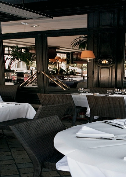 Een chique terras van een restaurant met ronde tafels, witte tafelkleden en keurig opgestelde glazen. De elegante, donkere inrichting en weelderige planten zorgen voor een luxueuze sfeer.