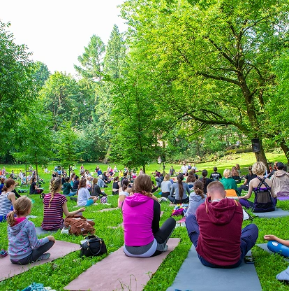 Een grote groep mensen neemt deel aan een yogales in de buitenlucht in een weelderig groen park. Ze zitten op matjes met hun gezicht naar de instructeur, onder een stralende zon.