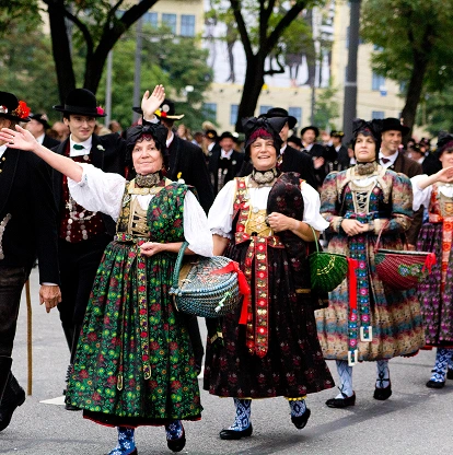 Een vrolijke optocht met mensen in traditionele Beierse klederdracht. Vrouwen in kleurrijke jurken en mannen in kostuums zwaaien in een feestelijke sfeer. Bomen en gebouwen vormen het decor.