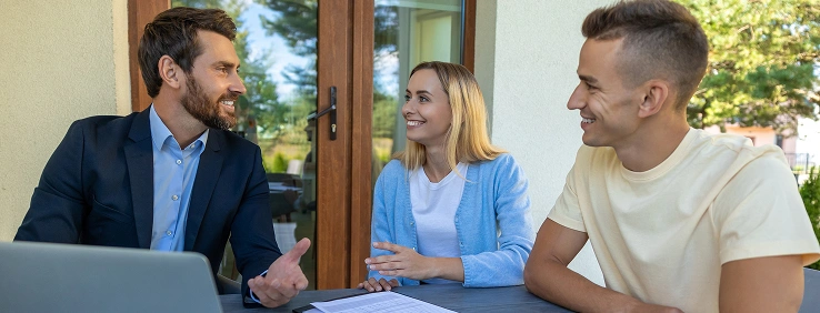 Een man in pak staat buiten te praten met een glimlachend stel. Op de tafel liggen een laptop en documenten. De sfeer is positief en er heerst een gevoel van samenwerking.